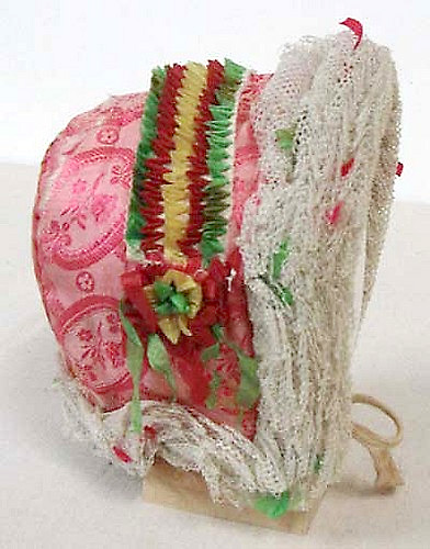 Kinderhaube (Chappin) aus rosaroter Seide mit weissem Taftband und farbigen Zierbändern, die zickzackartig aufgenäht sind.