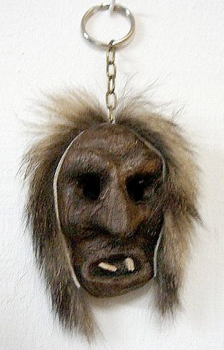Souvenirmaske als Schlüsselanhänger, 5 cm hohe Holzmaske mit eingesetzten Holzzähnen, umrandet von dünnem Fellstreifen.